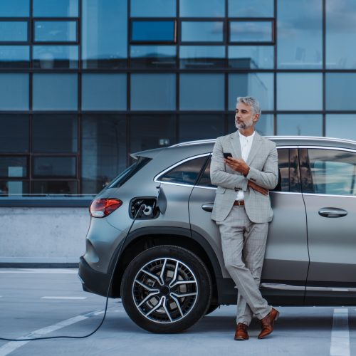 Businessman,Holding,Smartphone,While,Charging,Car,At,Electric,Vehicle,Charging Businessman holding smartphone while charging car at electric vehicle charging station, closeup.